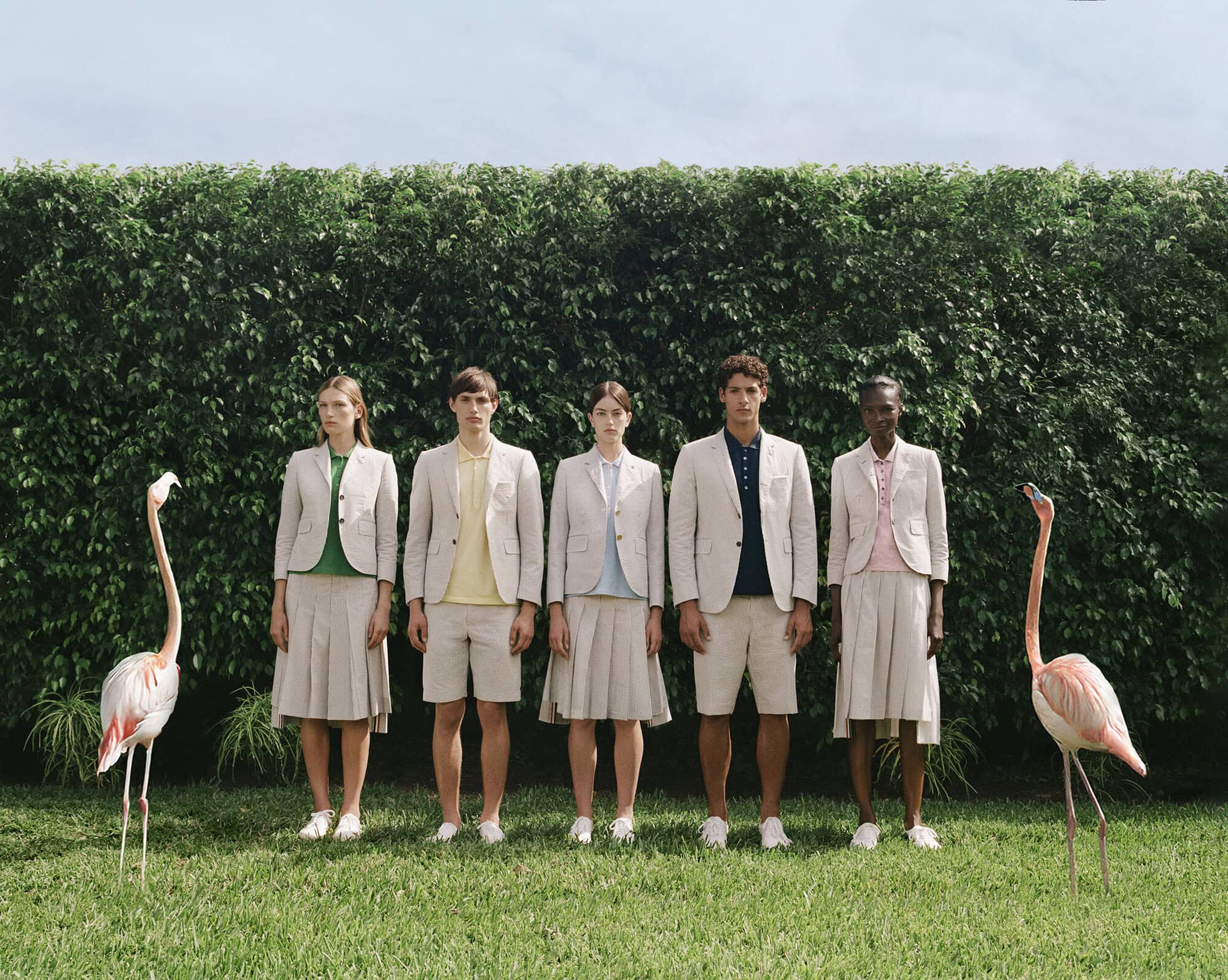 Five models in neutral linen suits standing symmetrically on a lawn flanked by two live flamingos before a tall green hedge.