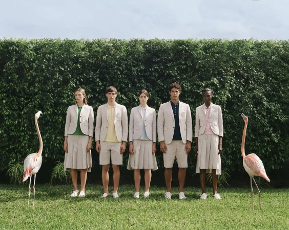 Five models in neutral linen suits standing symmetrically on a lawn flanked by two live flamingos before a tall green hedge.