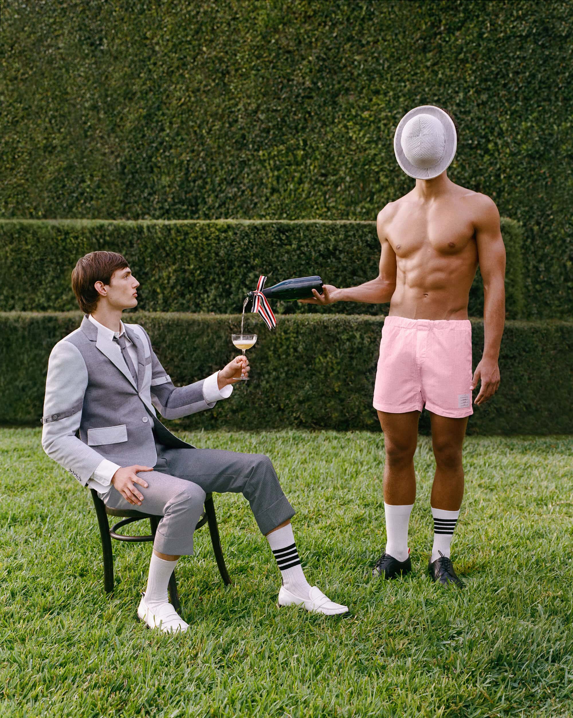 Seated man in a grey suit and a standing shirtless man holding a hat over his face while pouring a drink in a garden setting