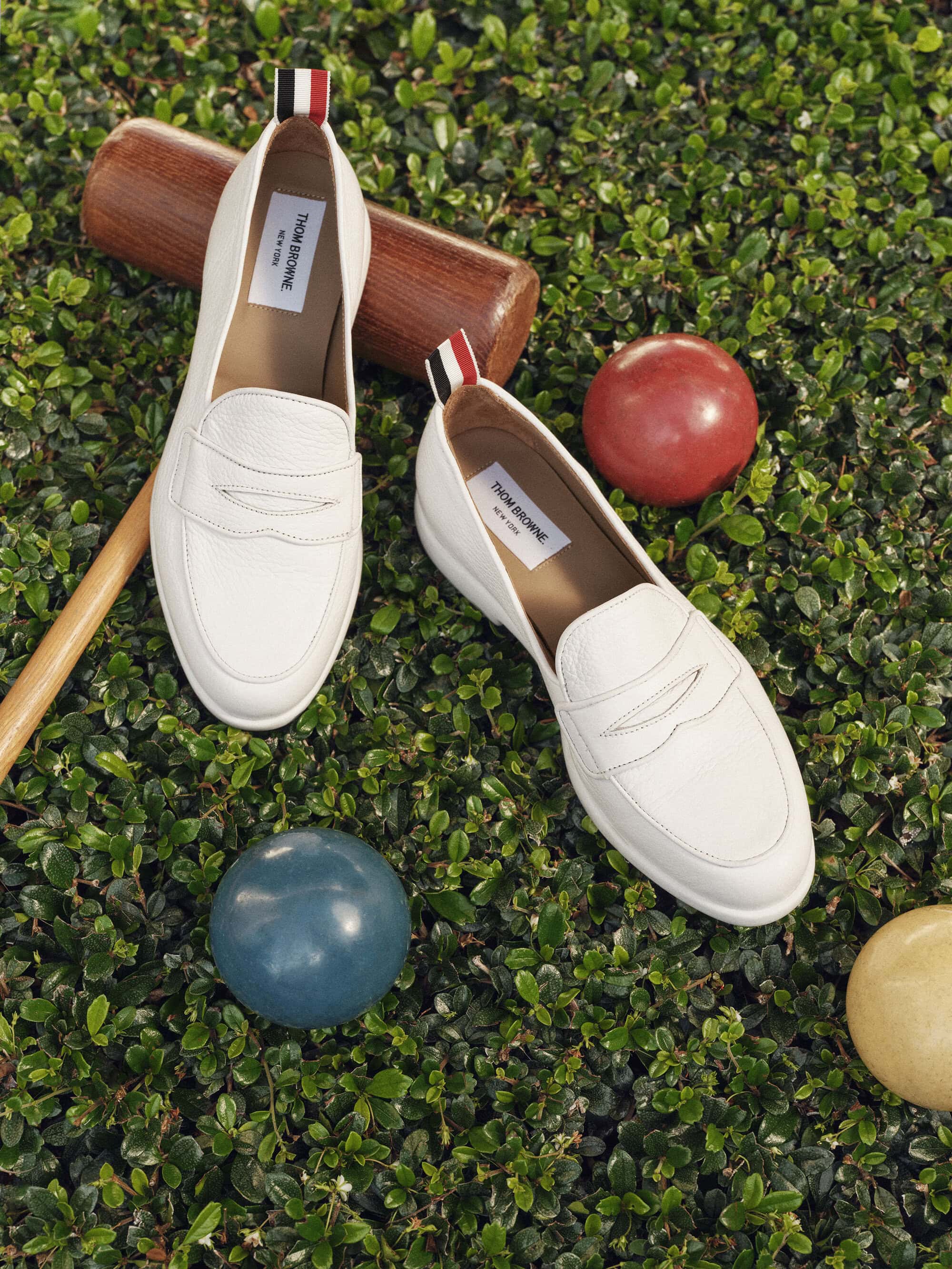 Two white leather penny loafers placed on lush green grass alongside croquet balls and a wooden mallet.