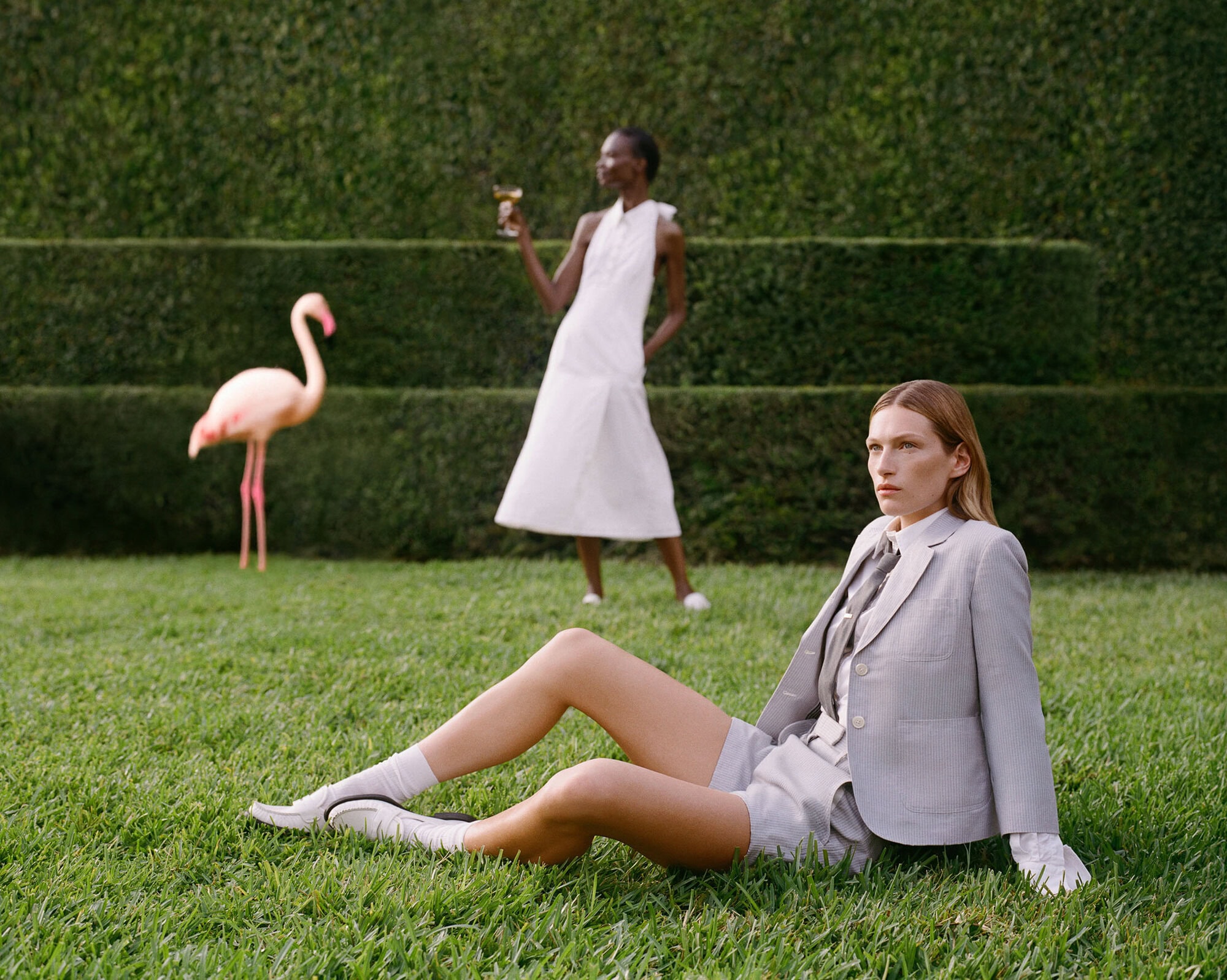 Two models on a manicured lawn with a pink flamingo statue, one seated in a light grey suit and the other standing in a white dress.