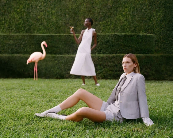 Two models on a manicured lawn with a pink flamingo statue, one seated in a light grey suit and the other standing in a white dress.