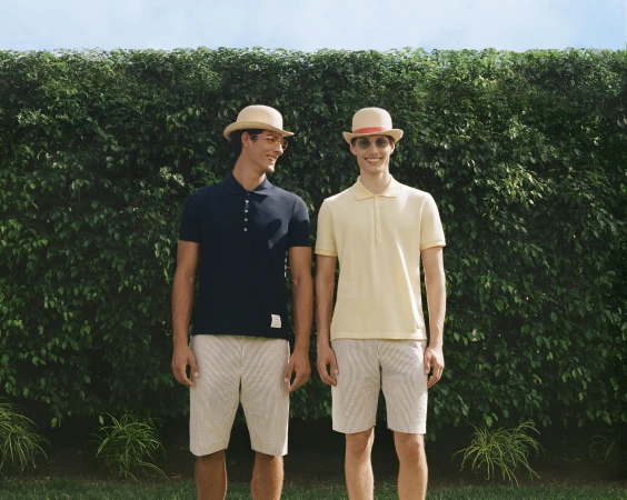 Two male models in polo shirts and bowler hats standing outdoors against a green hedge for Thom Browne summer campaign.