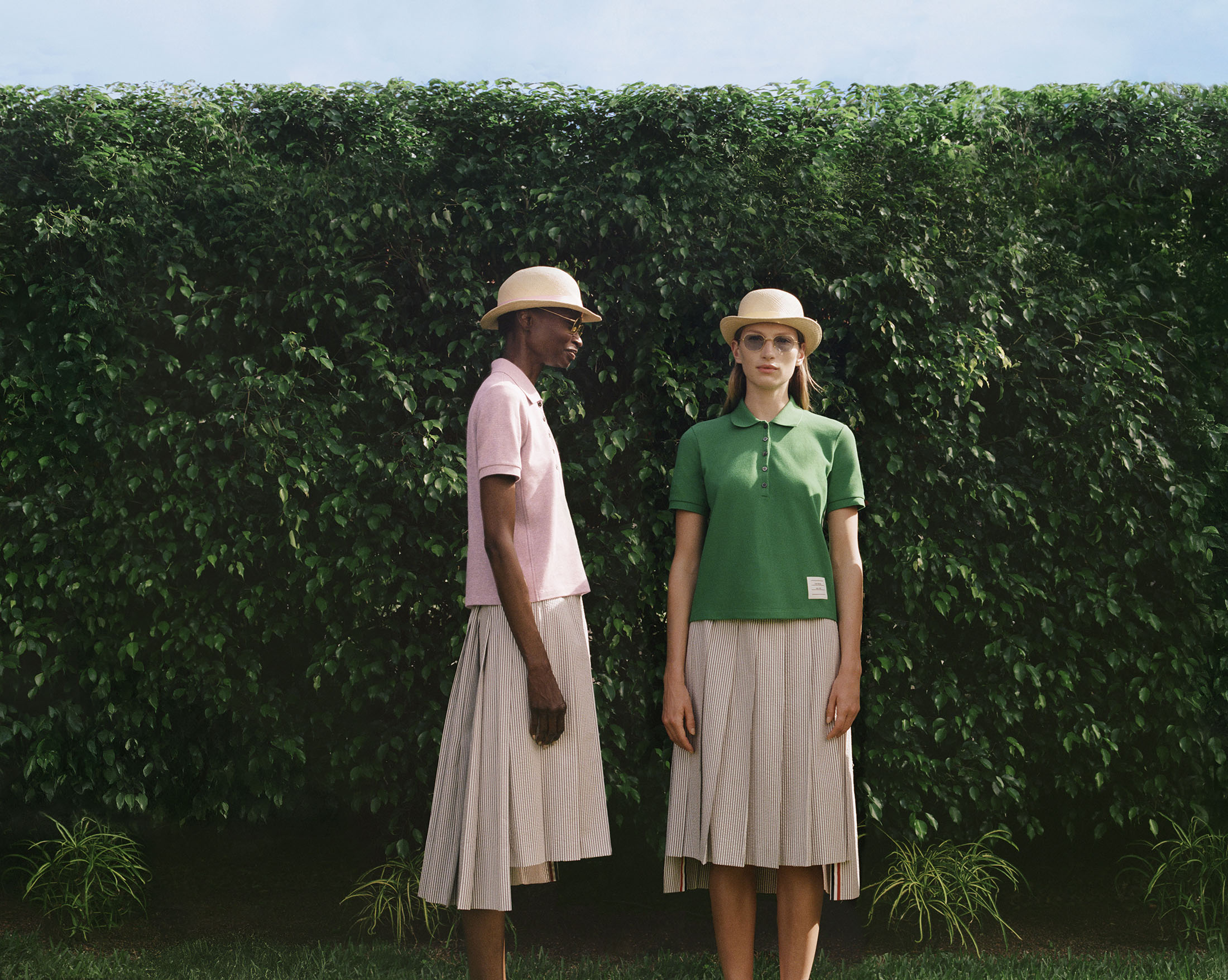 Two models wearing polo shirts, pleated skirts, and bowler hats standing in front of a dense green hedge.