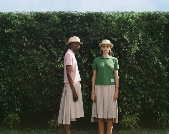 Two models wearing polo shirts, pleated skirts, and bowler hats standing in front of a dense green hedge.