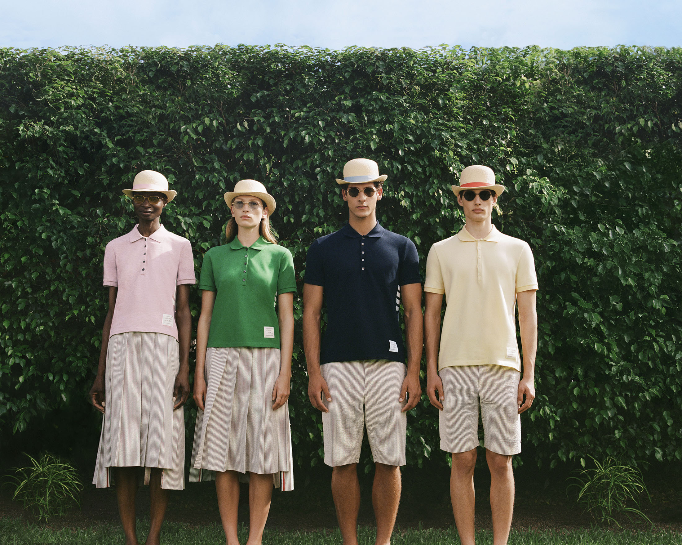 Four models wearing polo shirts, matching bowler hats, and sunglasses standing in a row against a tall green hedge.