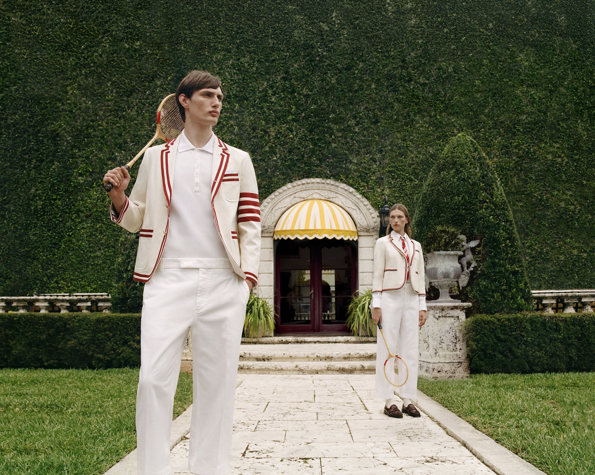 A man in a cream tennis blazer holding a racket stands in the foreground with a woman holding a racket in the background.