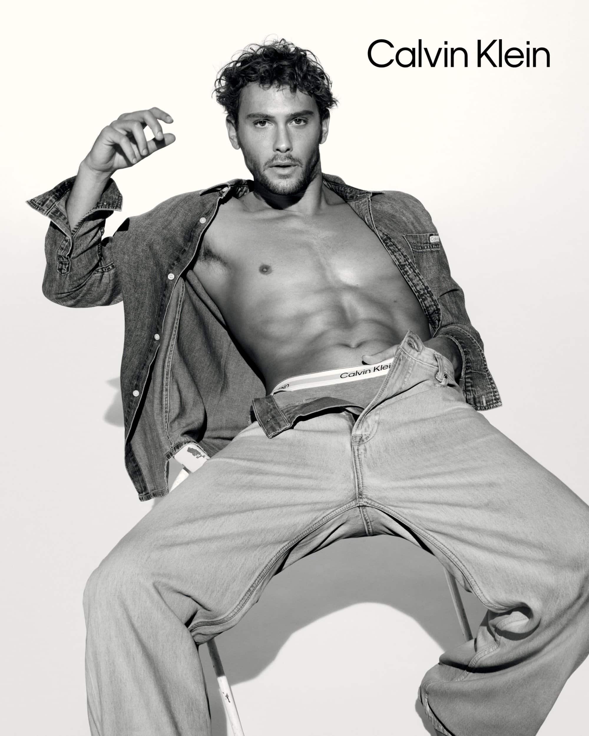 Model Cooper Koch seated wearing unbuttoned denim shirt and jeans for Calvin Klein spring 2025 ad campaign against white seamless background