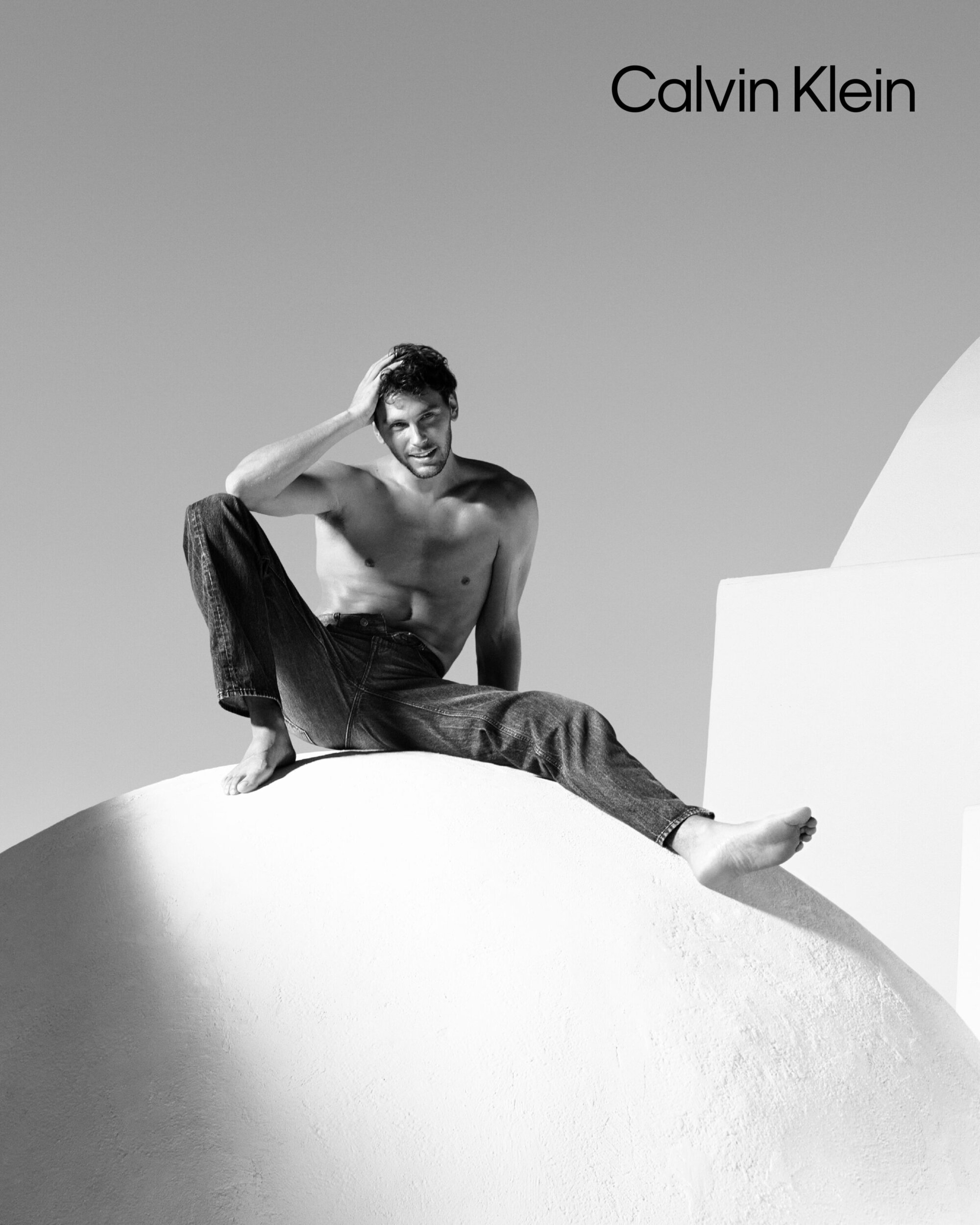 Model Cooper Koch sitting shirtless in jeans on a white architectural dome for Calvin Klein Spring 2025 ad campaign.