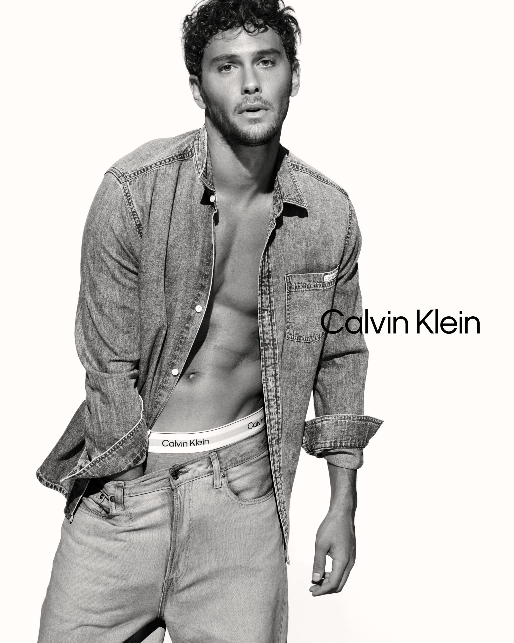 Cooper Koch posing in an unbuttoned denim shirt and jeans against a white seamless background for Calvin Klein.
