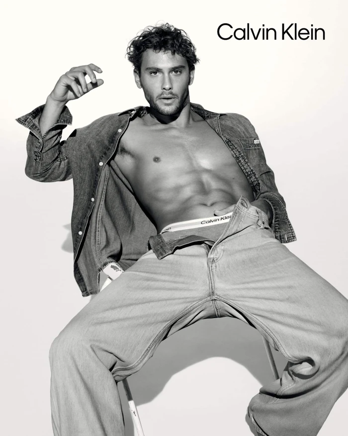Model Cooper Koch seated wearing unbuttoned denim shirt and jeans for Calvin Klein spring 2025 ad campaign against white seamless background
