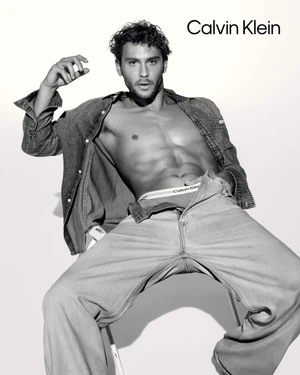 Model Cooper Koch seated wearing unbuttoned denim shirt and jeans for Calvin Klein spring 2025 ad campaign against white seamless background