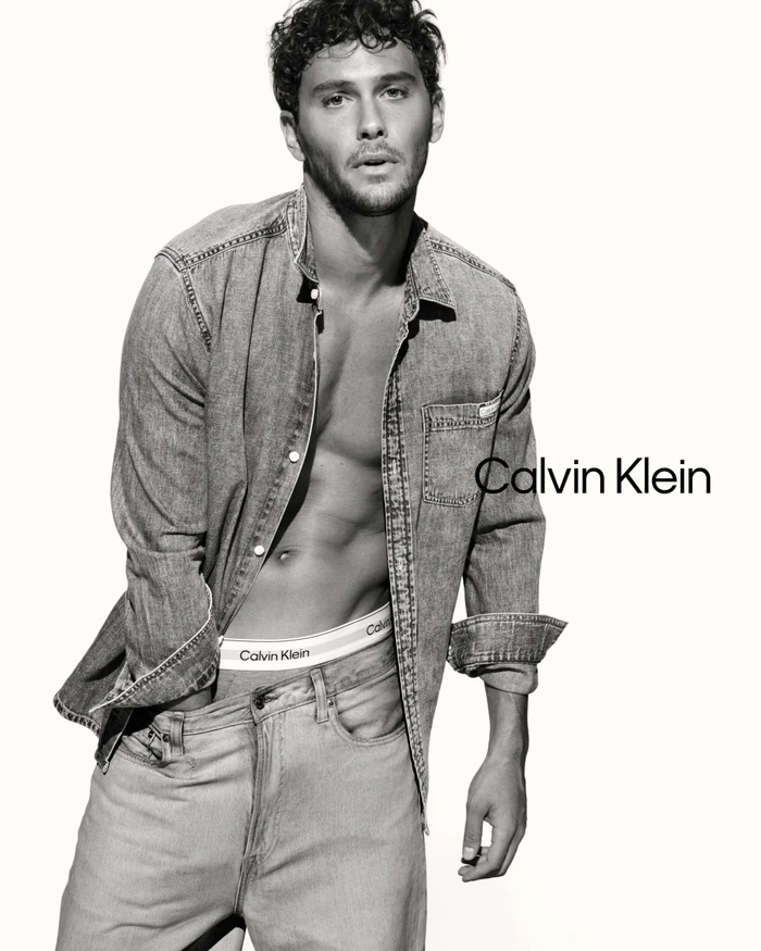 Cooper Koch posing in an unbuttoned denim shirt and jeans against a white seamless background for Calvin Klein.
