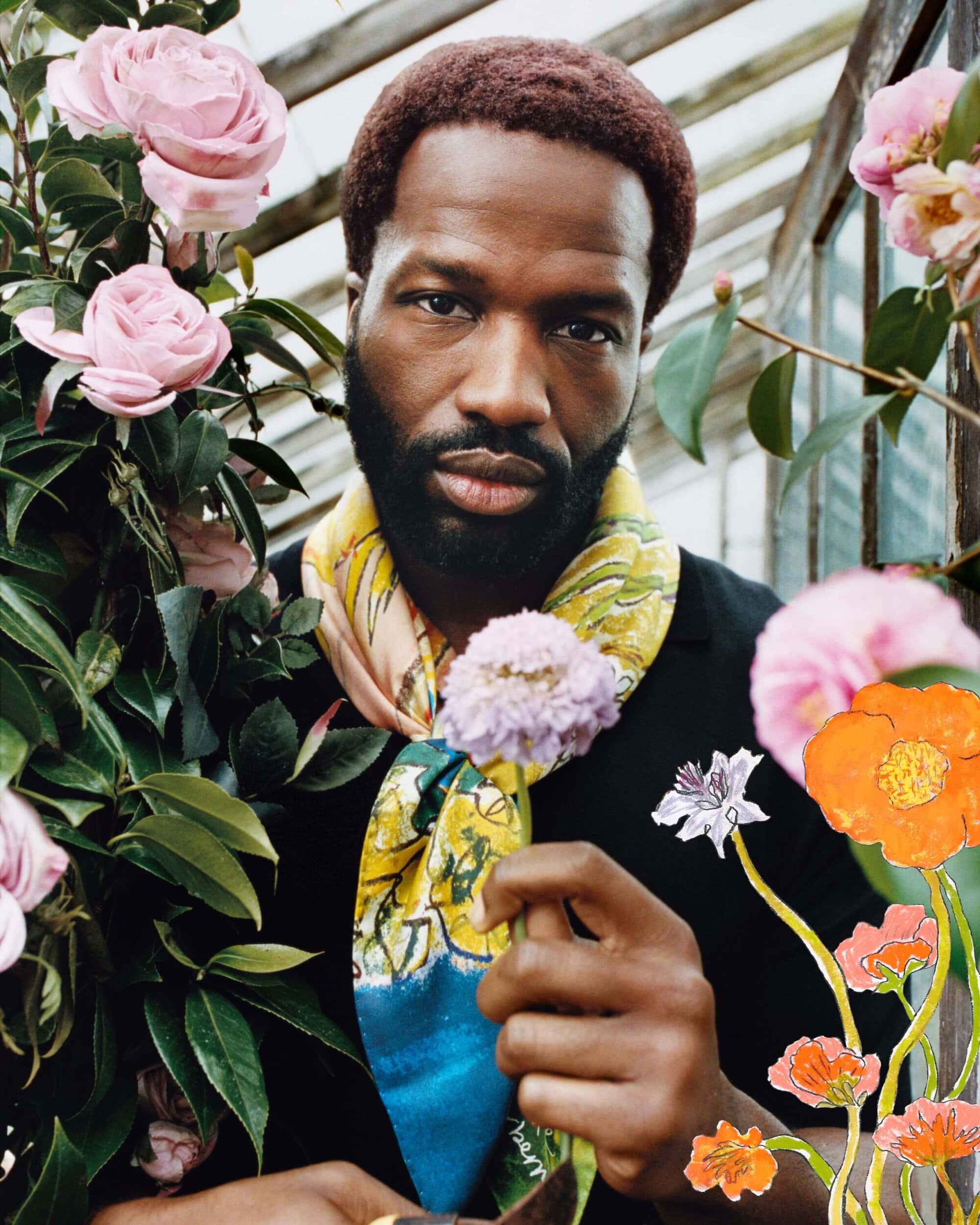 Black man holding a flower wearing a patterned scarf inside a greenhouse with roses