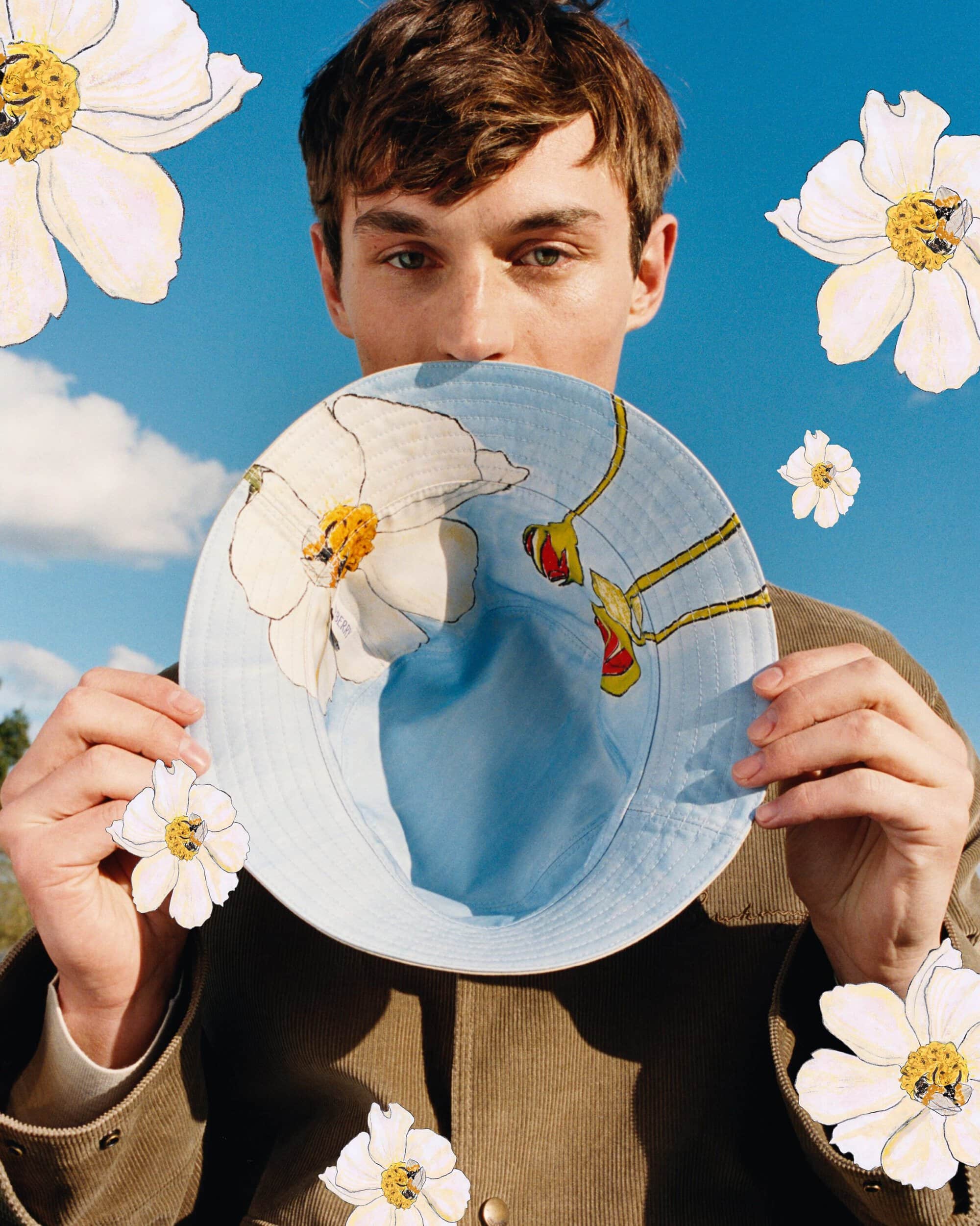 A man holding a painted blue bucket hat in front of his face beneath a clear blue sky, surrounded by illustrated white flowers.