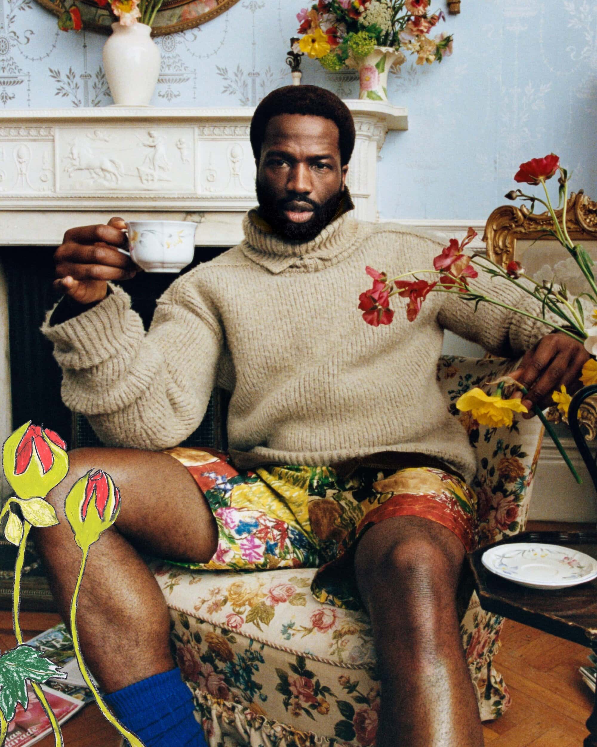 Man with beard seated in a floral armchair holding a teacup against a blue wallpaper interior background