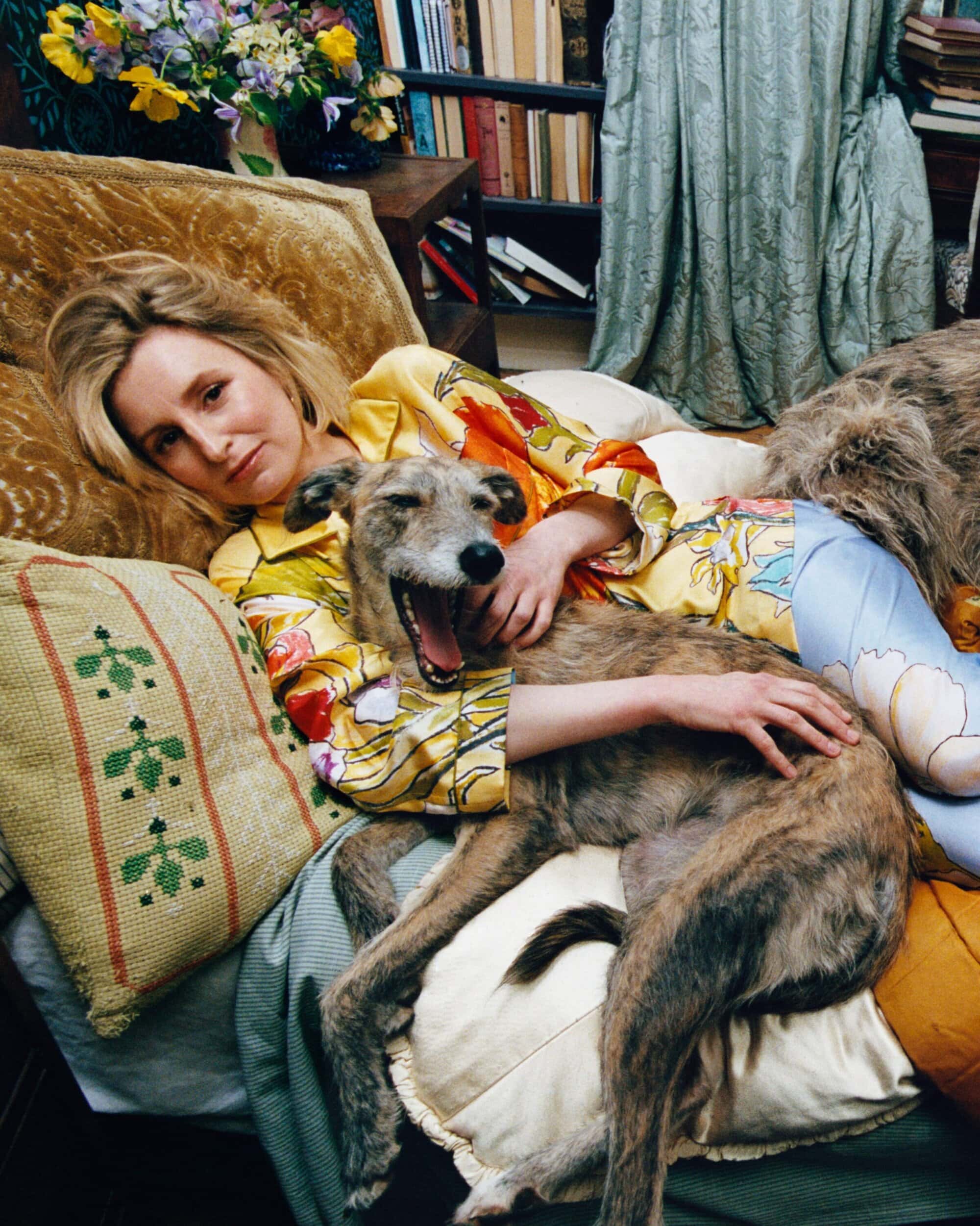 Woman lying on a sofa with a yawning dog, wearing a colorful patterned top in an eclectic room interior.