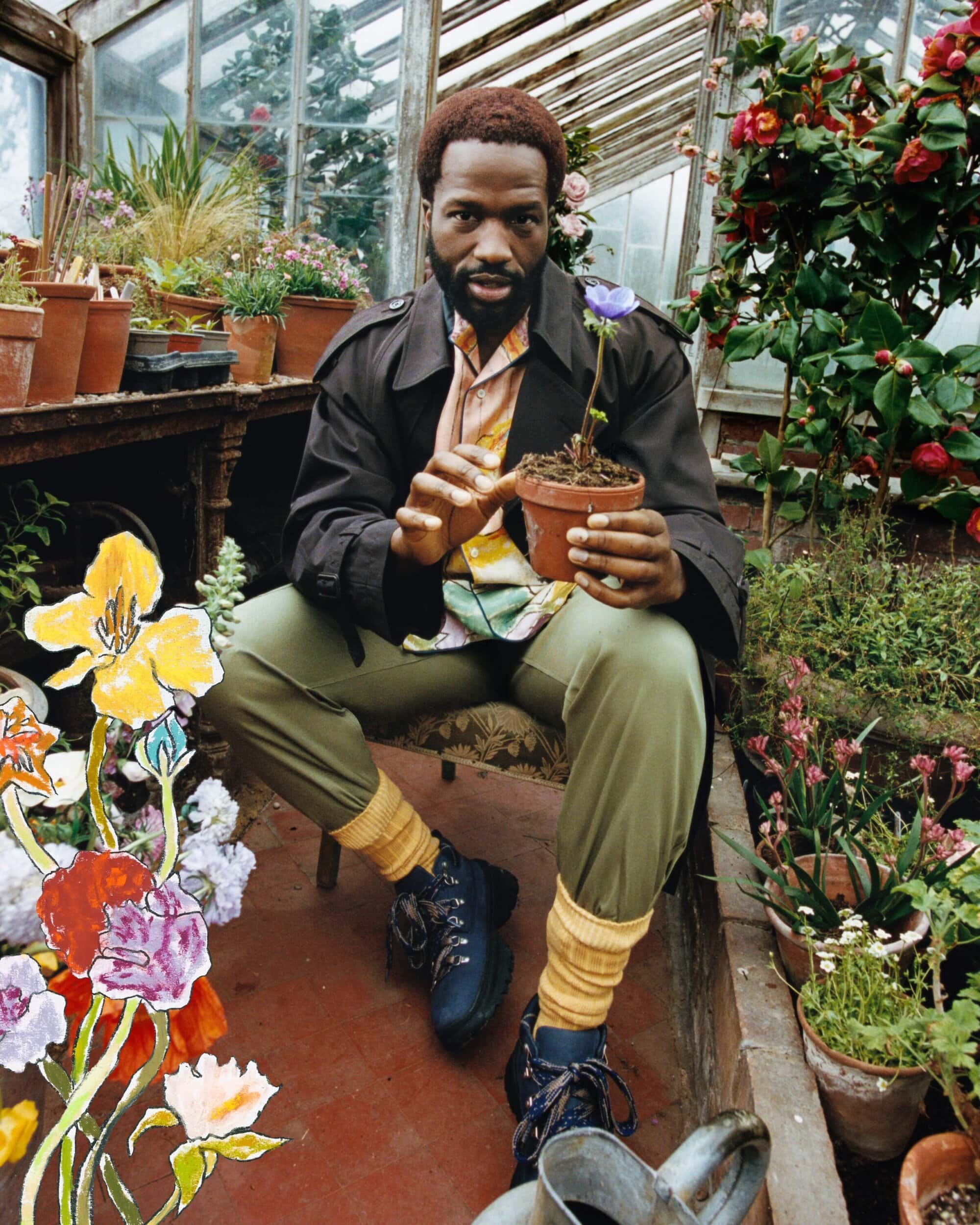 Black man holding a potted flower inside a greenhouse, wearing a brown coat and olive trousers with yellow socks.