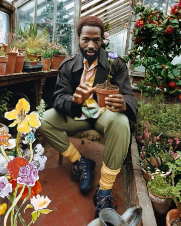 Black man holding a potted flower inside a greenhouse, wearing a brown coat and olive trousers with yellow socks.