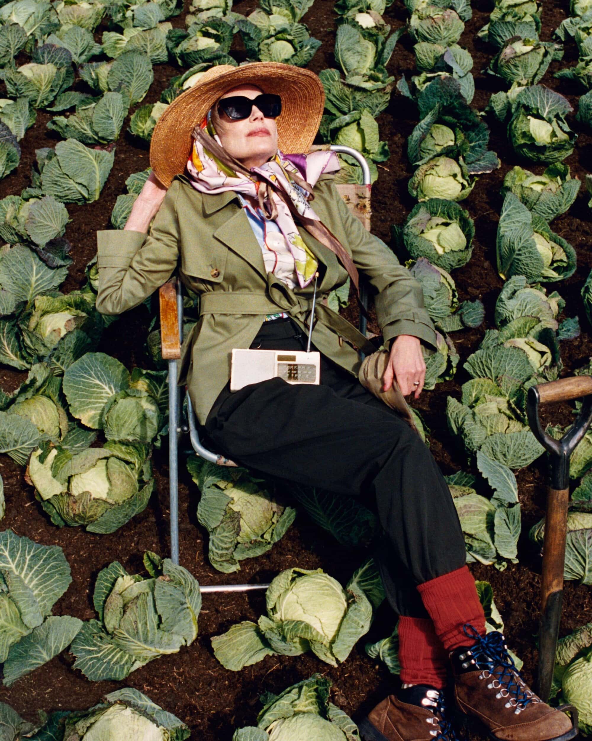 An older woman in a straw hat and green trench coat seated in a folding chair amidst a field of cabbages.