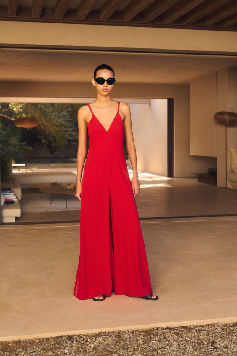 Fashionable woman in red palazzo jumpsuit and sunglasses stands on patio in front of modern neutral-toned home with open doorway.