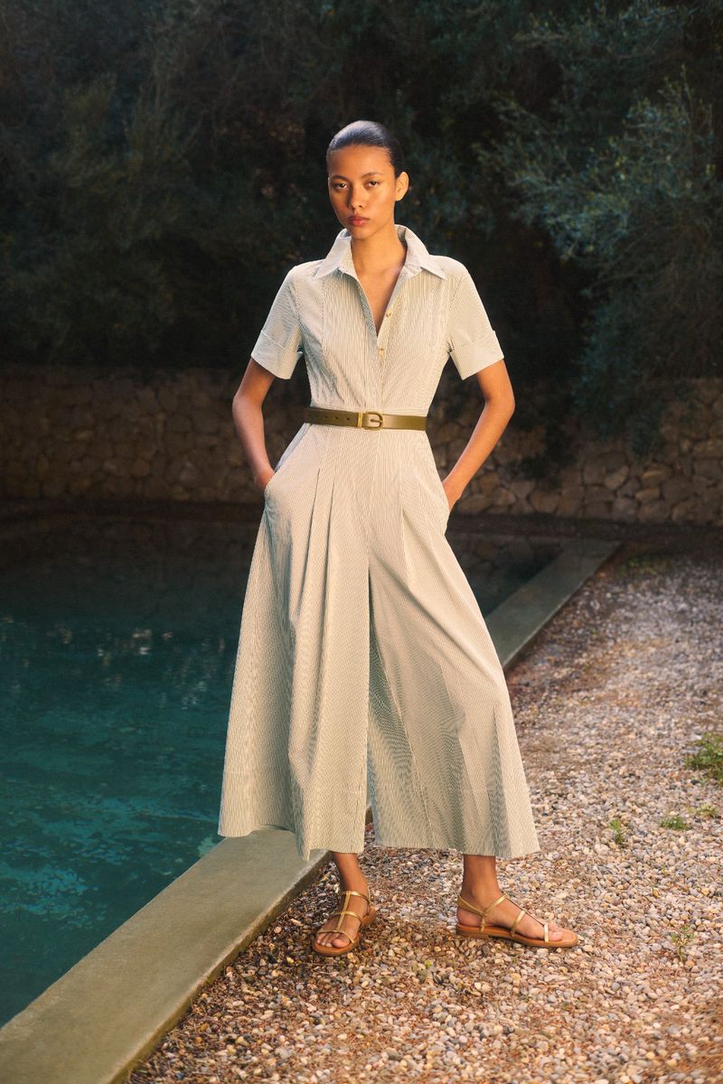 A woman in a striped jumpsuit poses outdoors near a pool. She's wearing gold sandals and a belt, standing on a gravel surface.