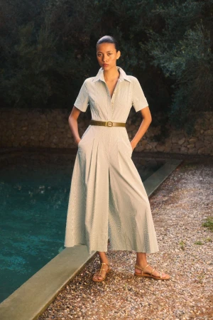 A woman in a striped jumpsuit poses outdoors near a pool. She's wearing gold sandals and a belt, standing on a gravel surface.