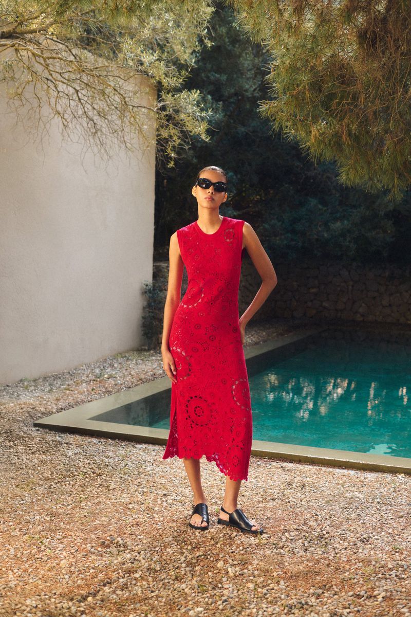 A fashionable woman stands near a pool wearing a red lace dress, sunglasses, and sandals, embodying a blend of modern and classic style.