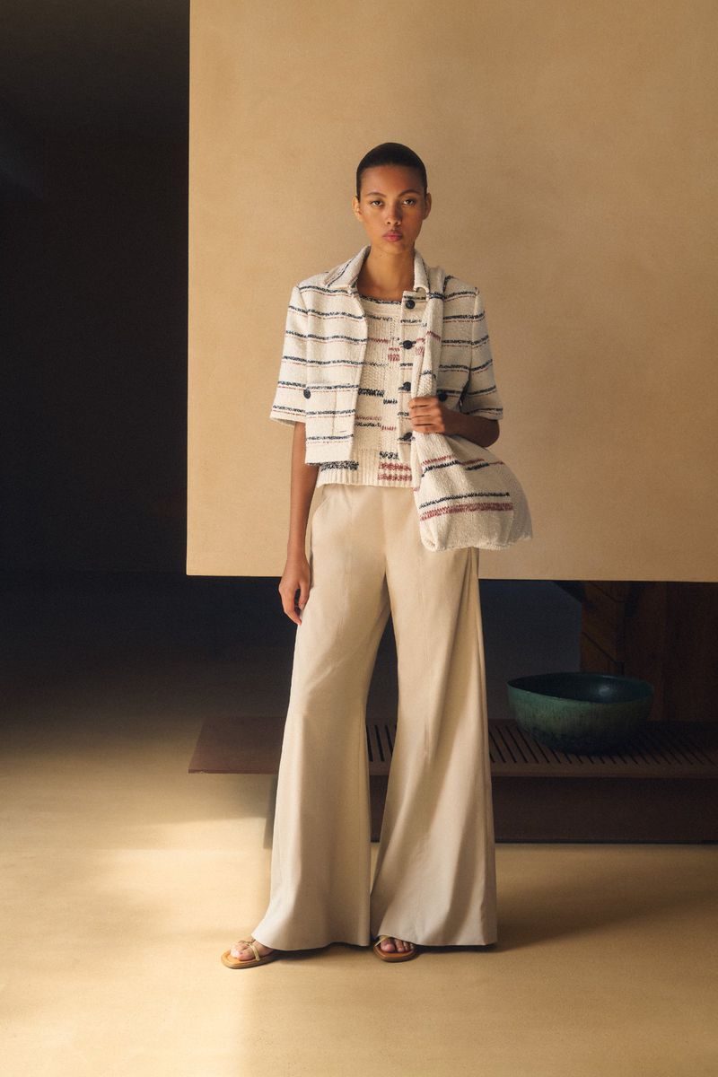 Fashion shot of a woman in a striped jacket and wide-leg pants, accessorized with a tote bag, standing in front of a neutral backdrop.