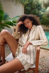 Model with curly hair seated in a wooden chair wearing a striped beige blazer and shorts outdoors by a pool