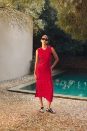 Model wearing a red sleeveless dress and black sunglasses standing outdoors near a swimming pool by a stone wall