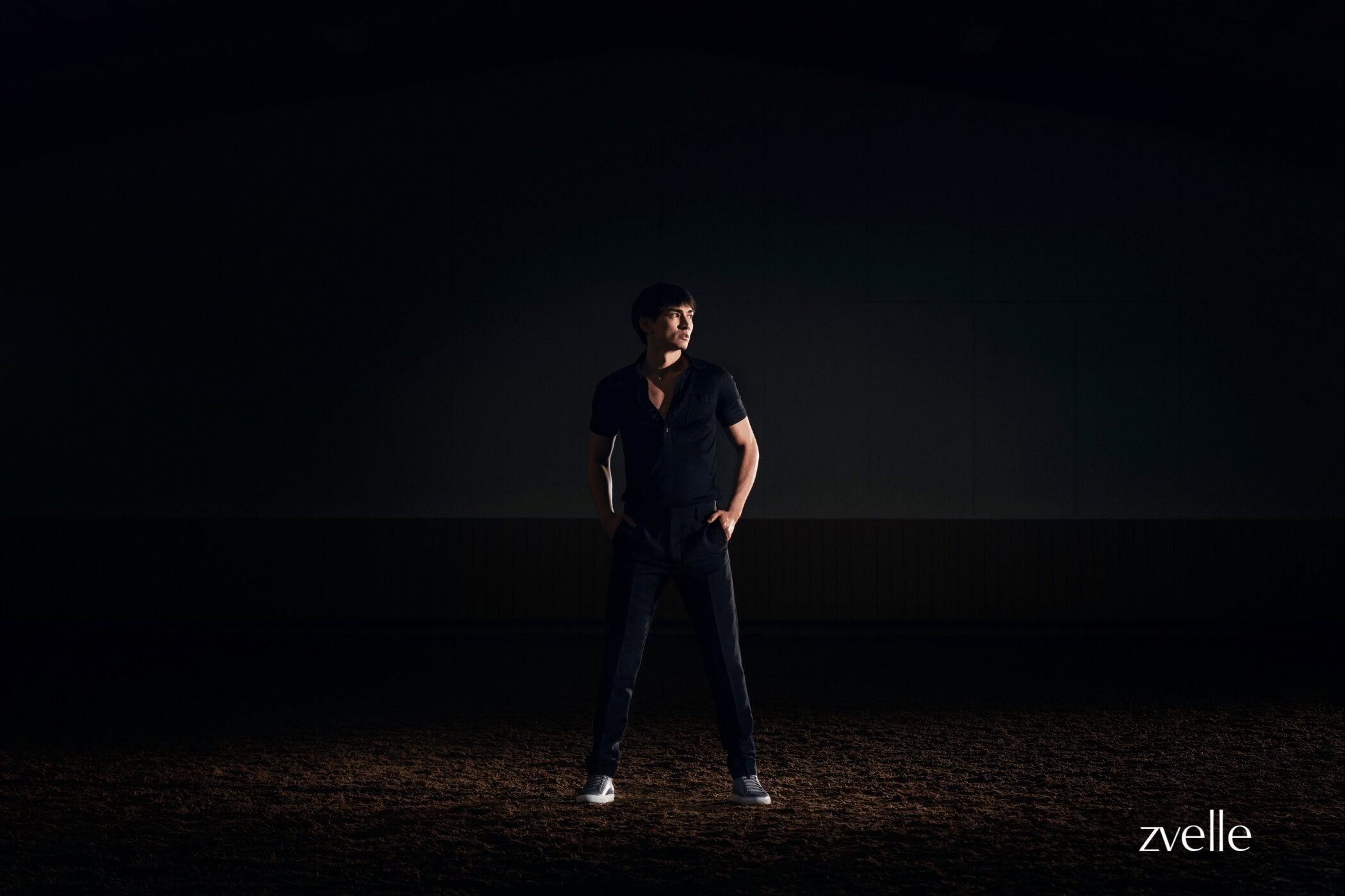 Man wearing a dark short-sleeve polo shirt and dark trousers standing on dark soil under dramatic spotlight.
