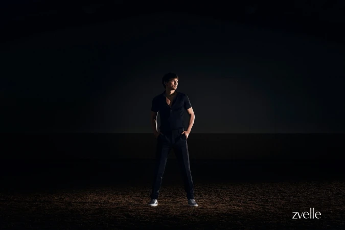 Man wearing a dark short-sleeve polo shirt and dark trousers standing on dark soil under dramatic spotlight.