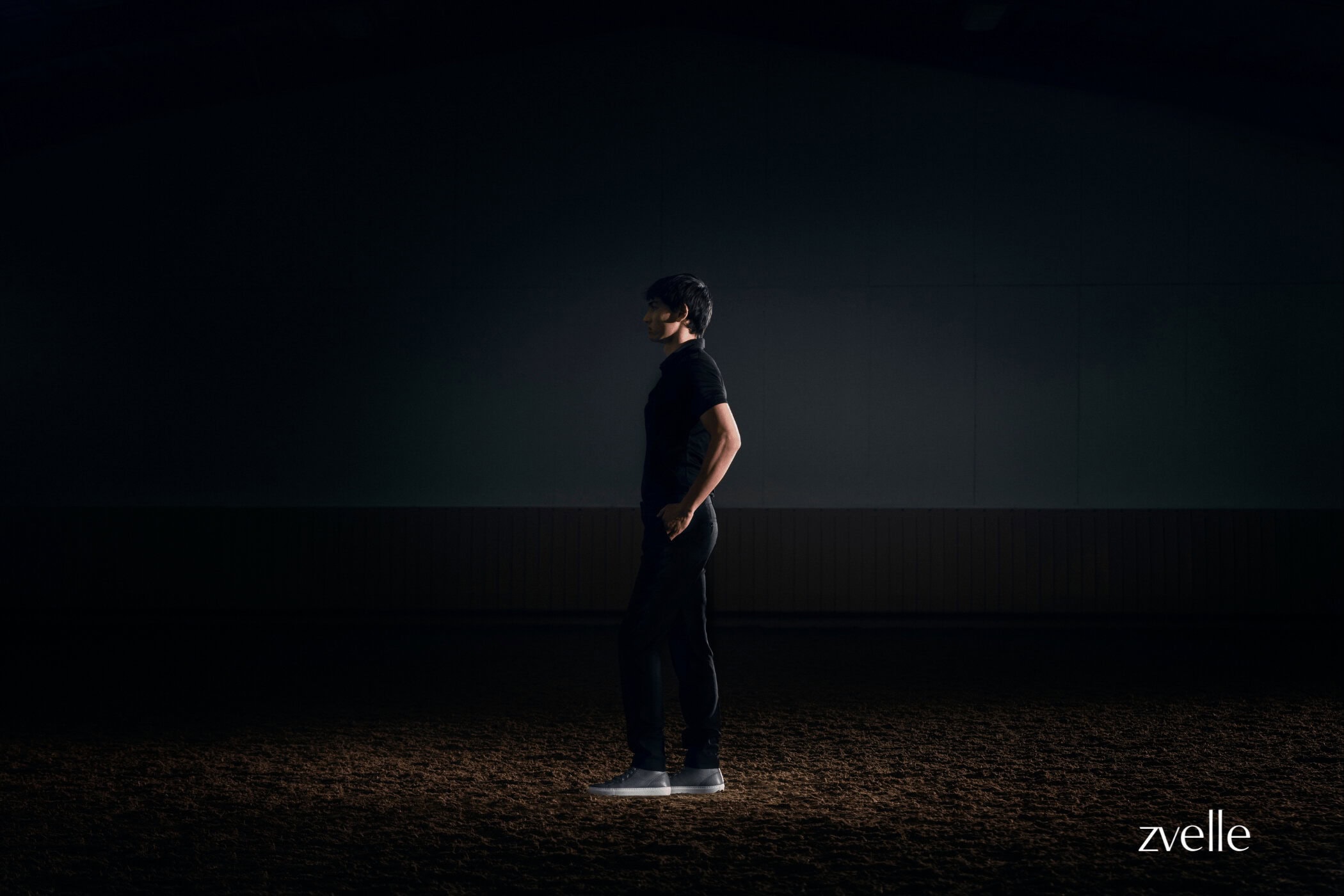 A person standing in profile in low-key lighting wearing dark clothing against a dark background for Zvelle spring 2025 ad campaign.