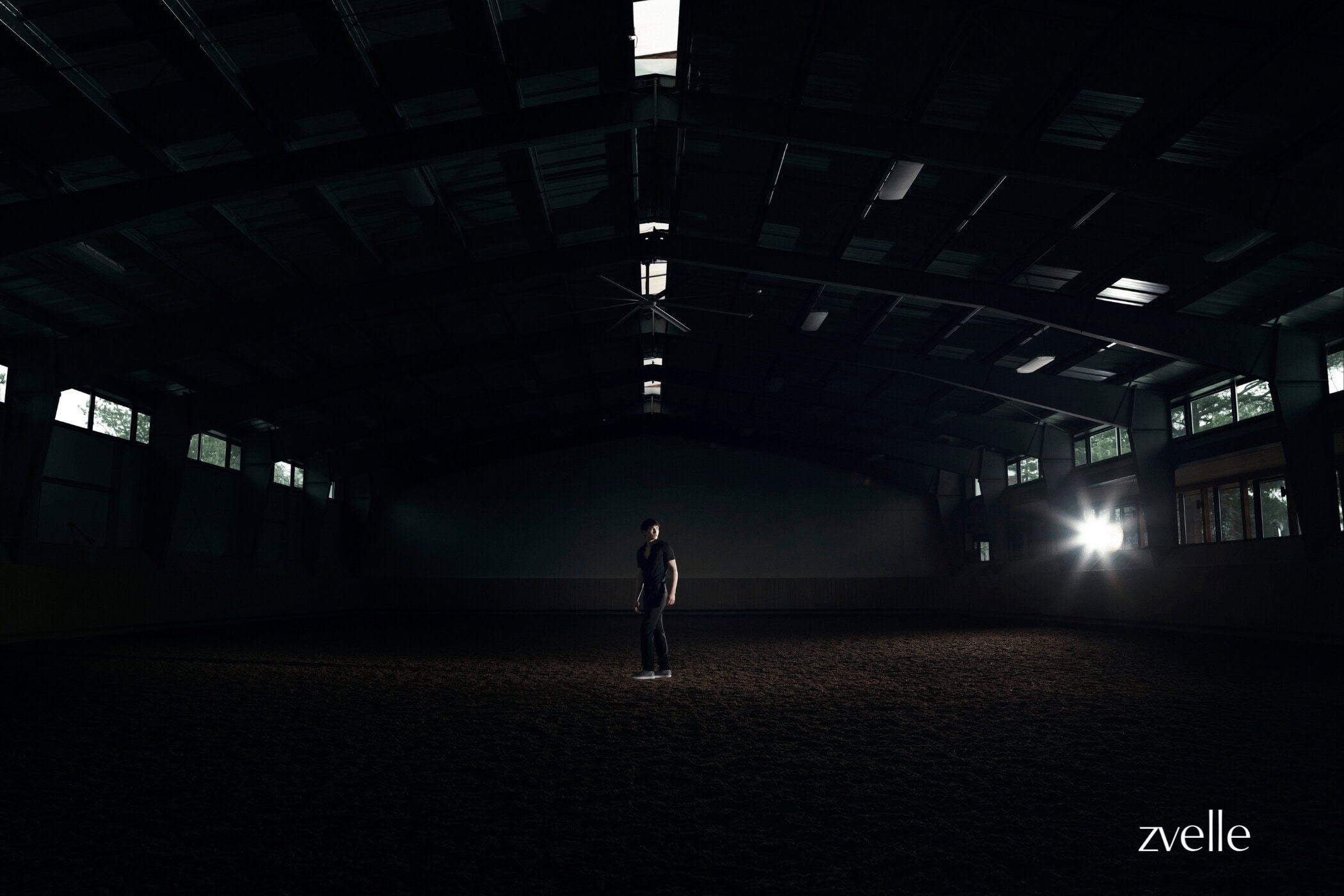 A person standing alone in the center of a large, dark indoor arena lit by a bright light source on the right.