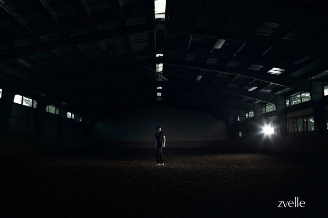 A person standing alone in the center of a large, dark indoor arena lit by a bright light source on the right.