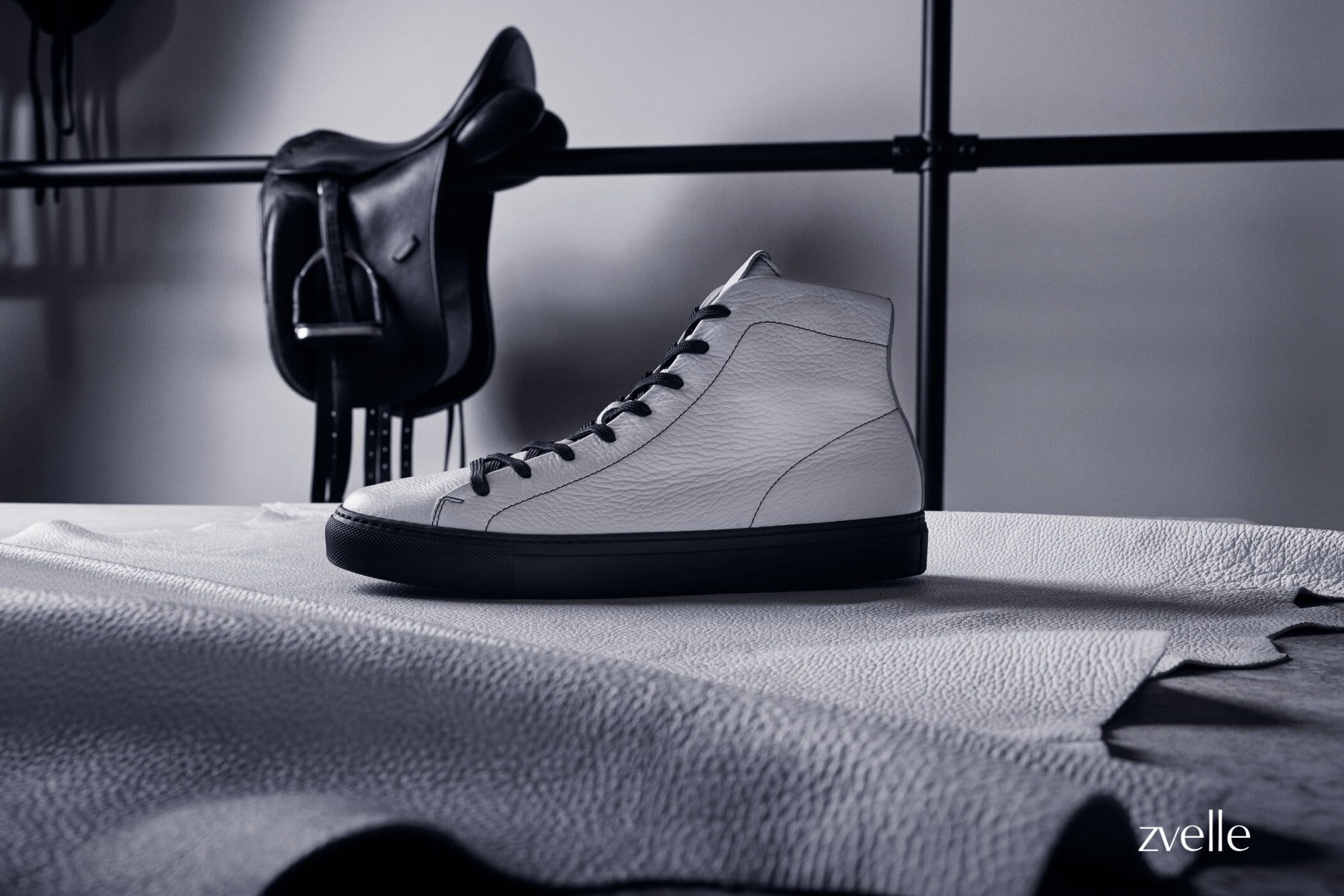White high top leather sneaker with dark laces resting on a textured leather sheet with a black saddle hanging in the background.