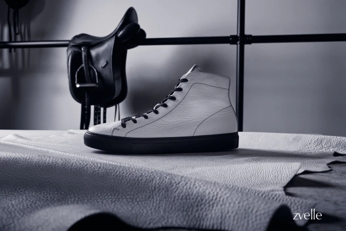 White high top leather sneaker with dark laces resting on a textured leather sheet with a black saddle hanging in the background.