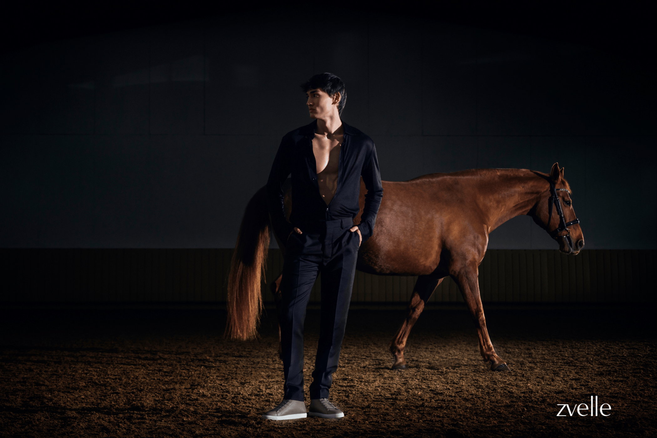 Man wearing dark shirt and trousers standing beside a brown horse on a dirt floor under low key lighting