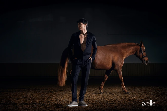Man wearing dark shirt and trousers standing beside a brown horse on a dirt floor under low key lighting
