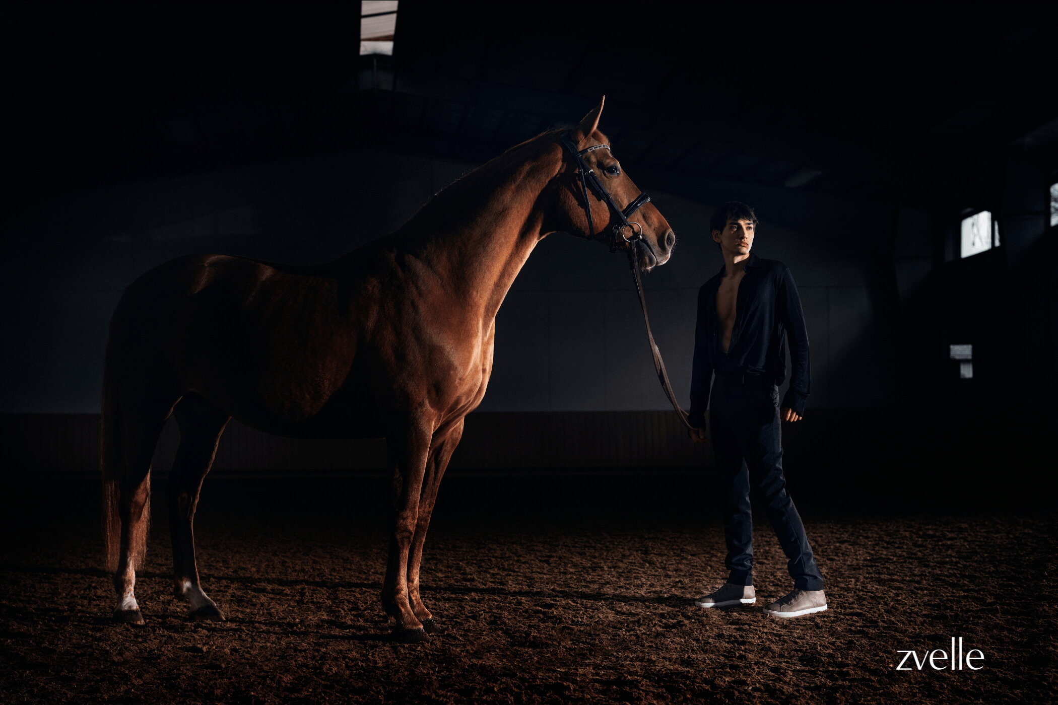 A man holding the reins of a brown horse standing in a dimly lit indoor arena for the Zvelle Spring 2025 ad campaign.