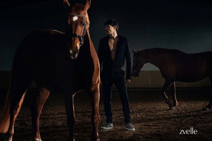 Man in black standing between two brown horses in an indoor arena for Zvelle spring 2025 ad campaign photographed by Daniel Harrison