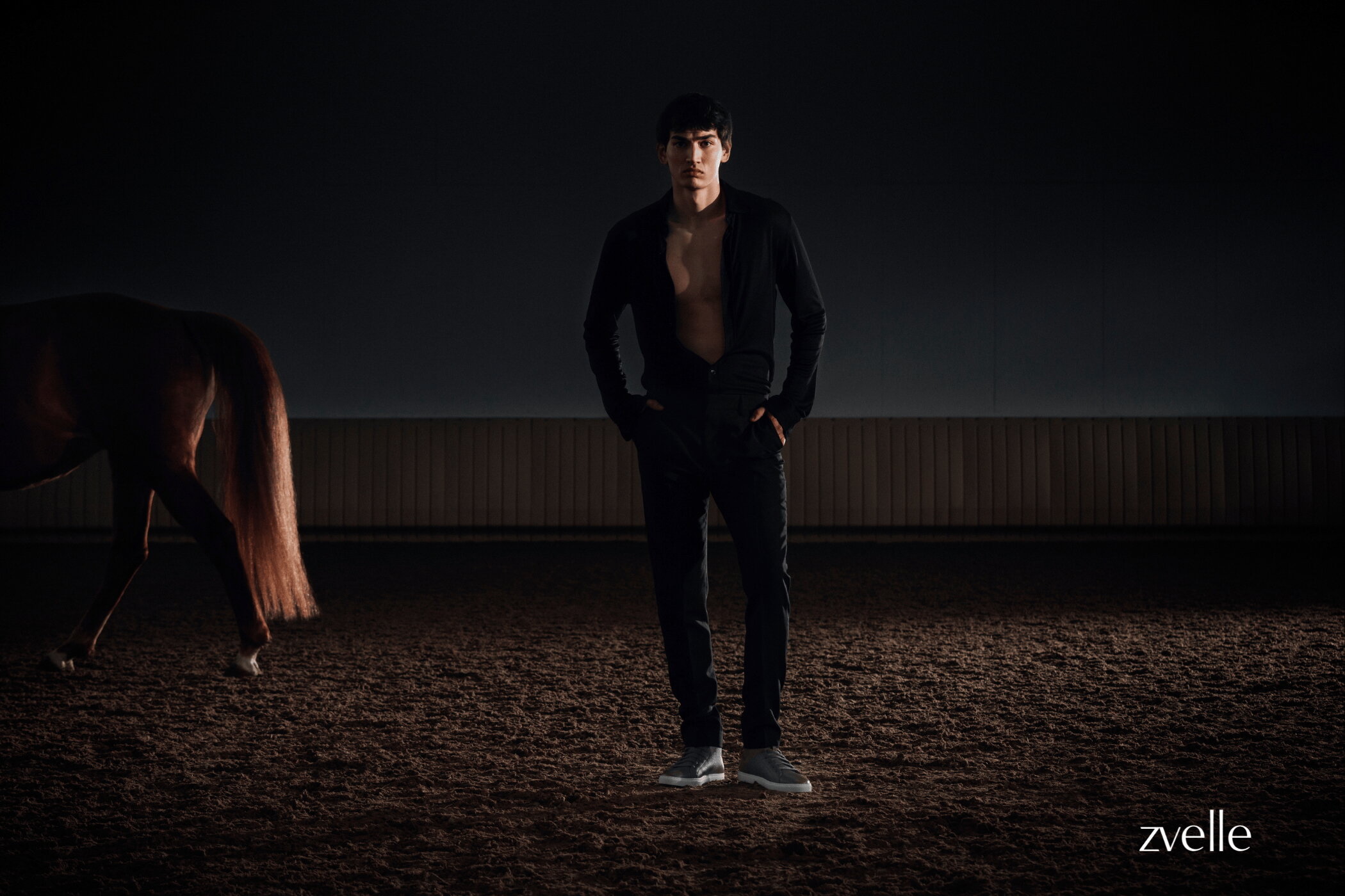 Man wearing a black shirt standing on a dirt ground next to a horse with a dark background