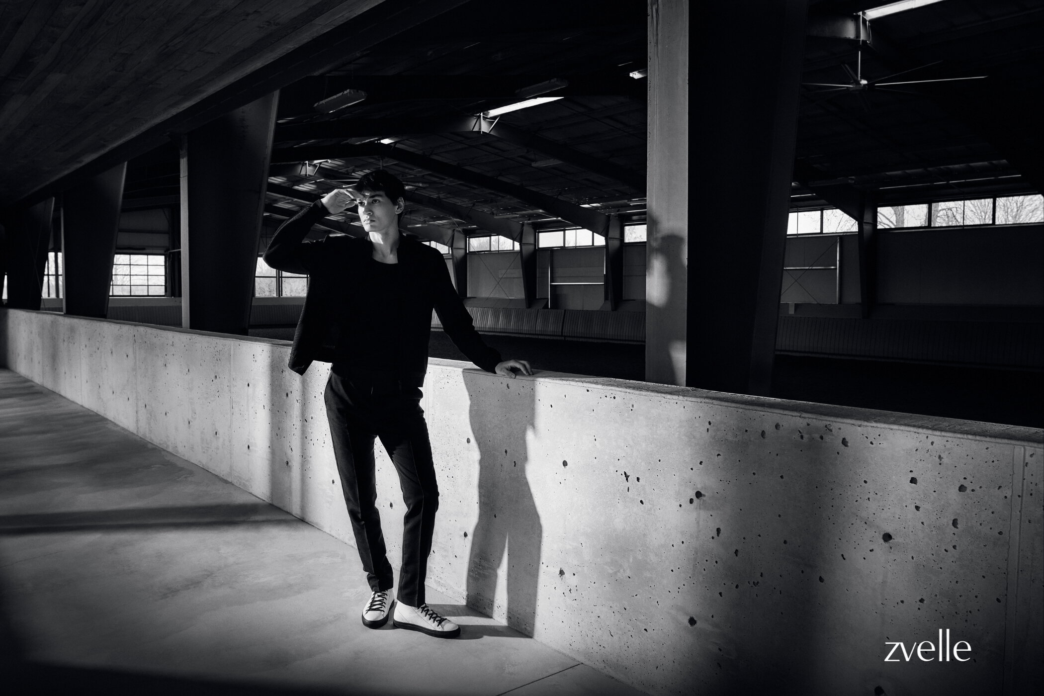 Man wearing dark jacket and trousers leaning against a concrete wall inside an industrial building interior