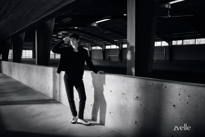 Man wearing dark jacket and trousers leaning against a concrete wall inside an industrial building interior