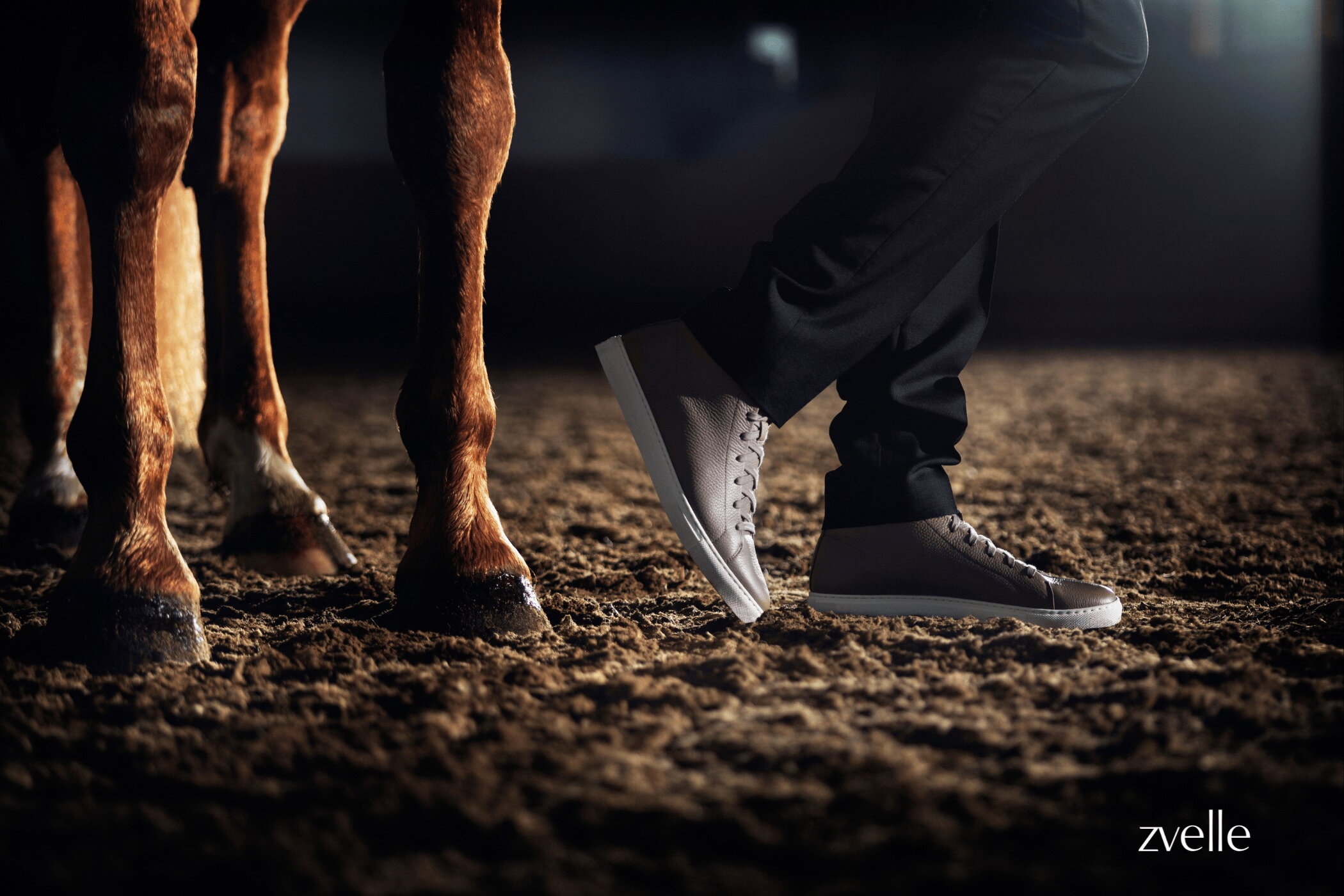 Close-up of a person wearing leather sneakers standing beside a horse in a stable arena, captured for Zvelle's Spring 2025 campaign.