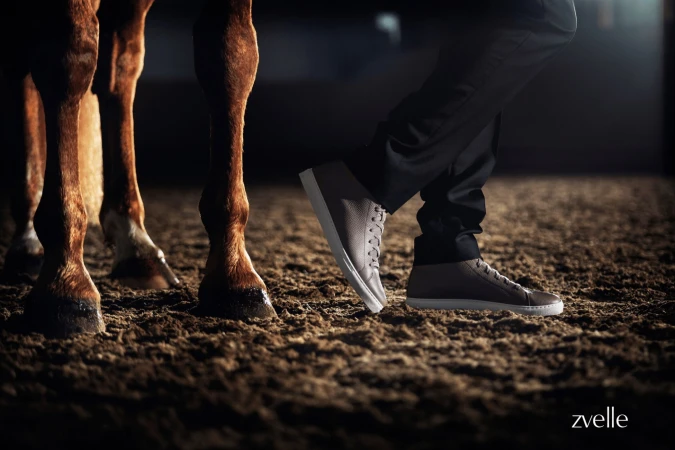 Close-up of a person wearing leather sneakers standing beside a horse in a stable arena, captured for Zvelle's Spring 2025 campaign.