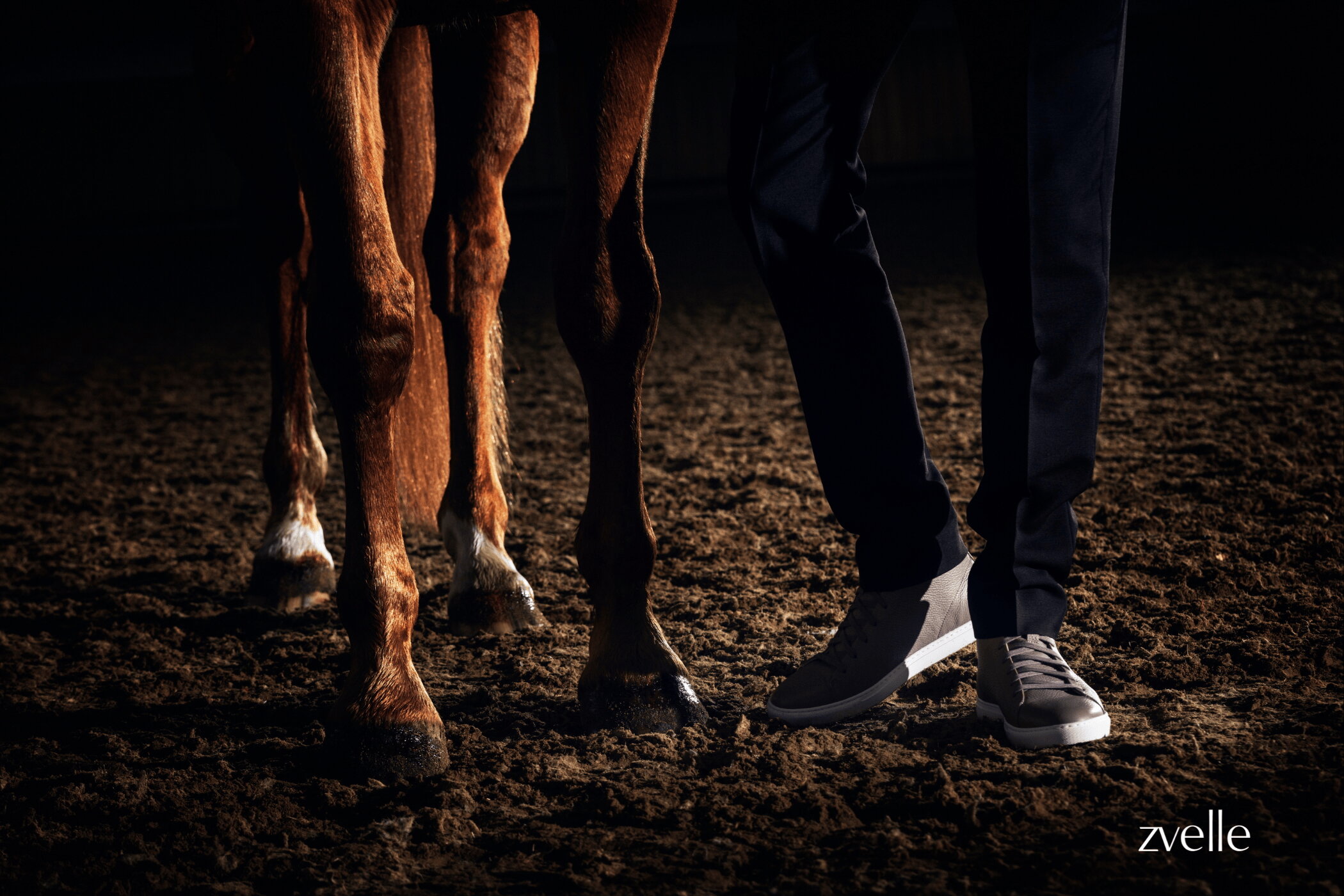 Low-angle shot focusing on a person's legs wearing grey sneakers and dark trousers standing beside a horse on a dirt surface.