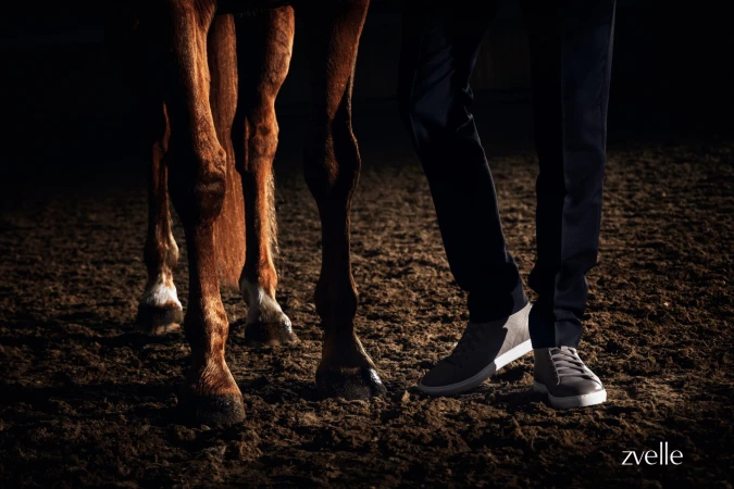 Low-angle shot focusing on a person's legs wearing grey sneakers and dark trousers standing beside a horse on a dirt surface.