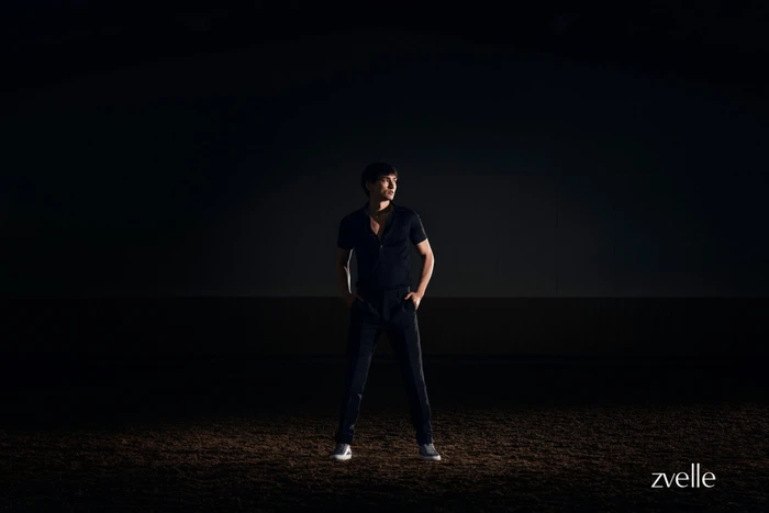 Man wearing a dark short-sleeve polo shirt and dark trousers standing on dark soil under dramatic spotlight.